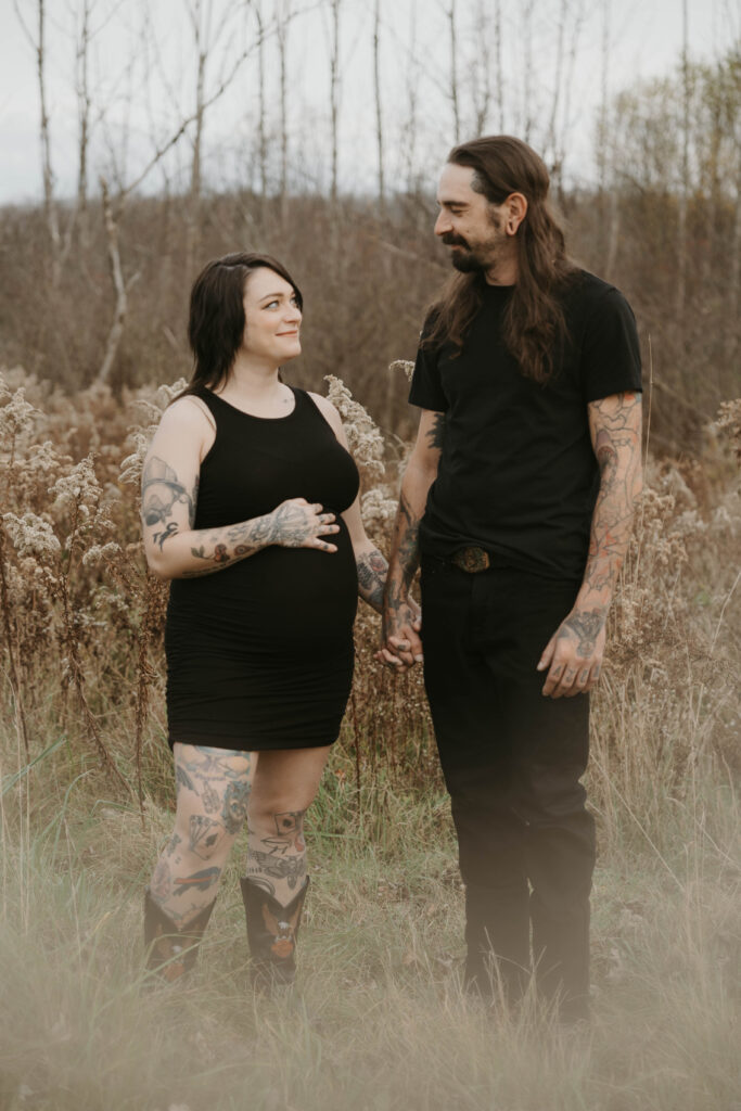 A slow moment of two soon-to-be parents in a field, they are holding hands and looking into eachothers eyes.