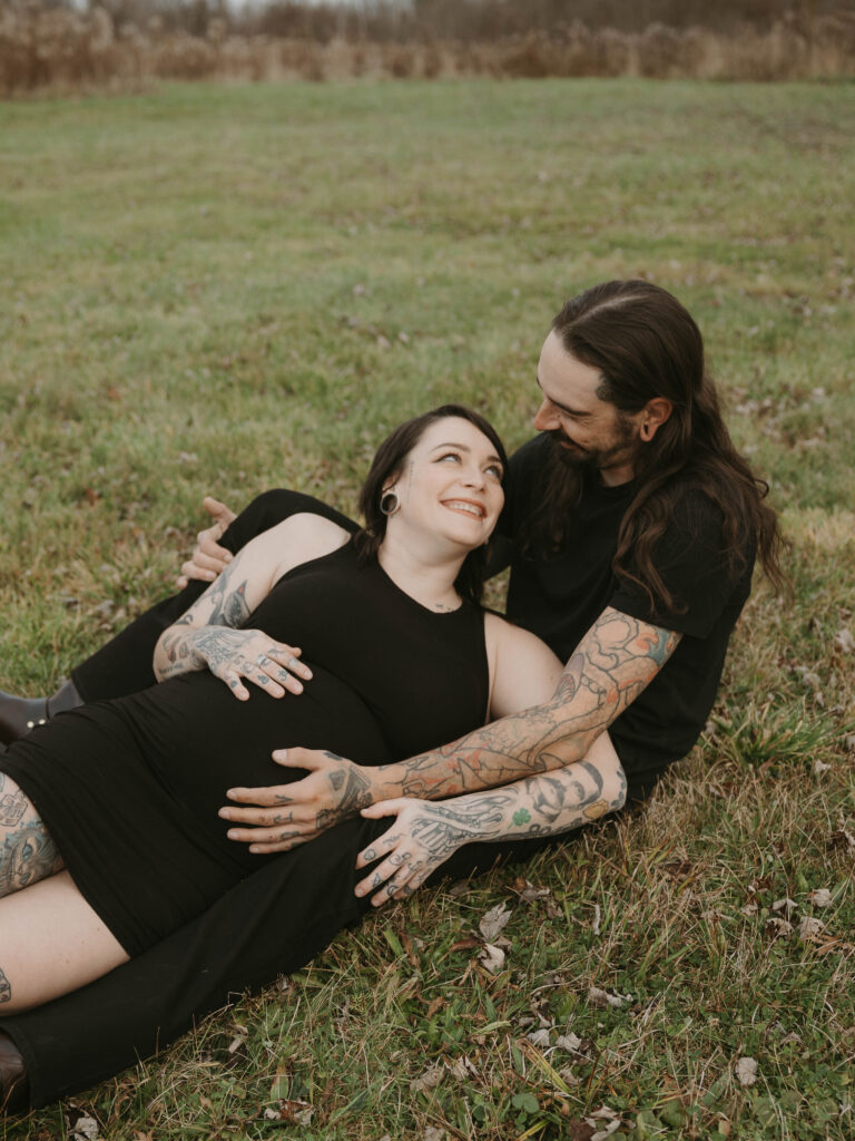An image of two soon-to-be parents laying in the grass, they laugh and smile as they embrace the growing belly.